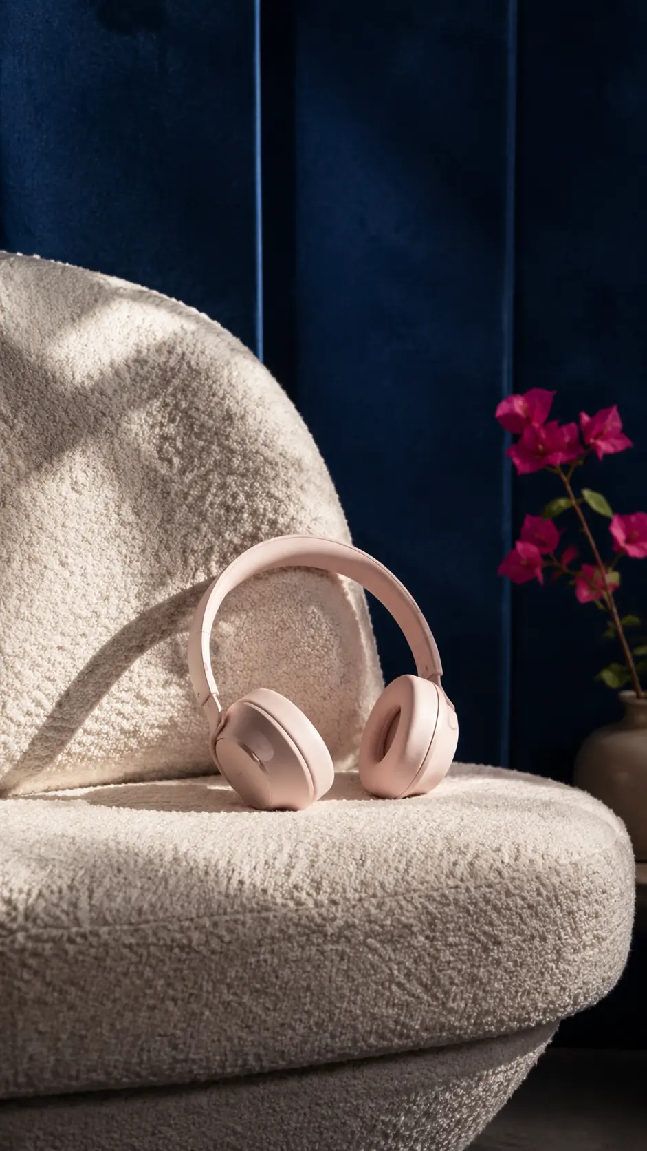 Blush headphones resting naturally on an ivory bouclé chair beside restrained fuchsia flowers