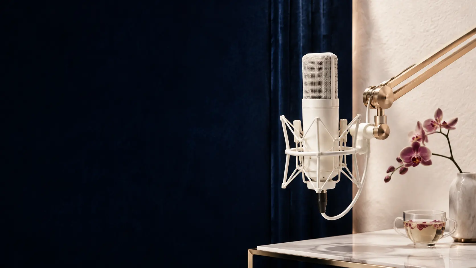 A pearl-white studio microphone beside botanical tea and an orchid in an ivory-and-navy recording space